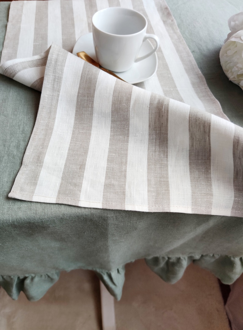 May include: A white and beige striped linen table runner with a ruffled green tablecloth underneath. A white coffee cup and saucer are on top of the table runner.