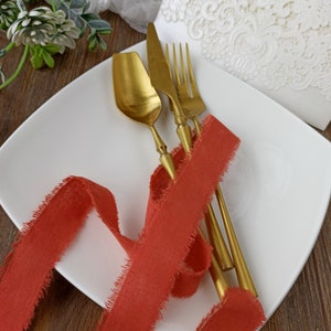 May include: A white plate with a red ribbon and gold cutlery. The ribbon has frayed edges and the cutlery is arranged on top of the ribbon.