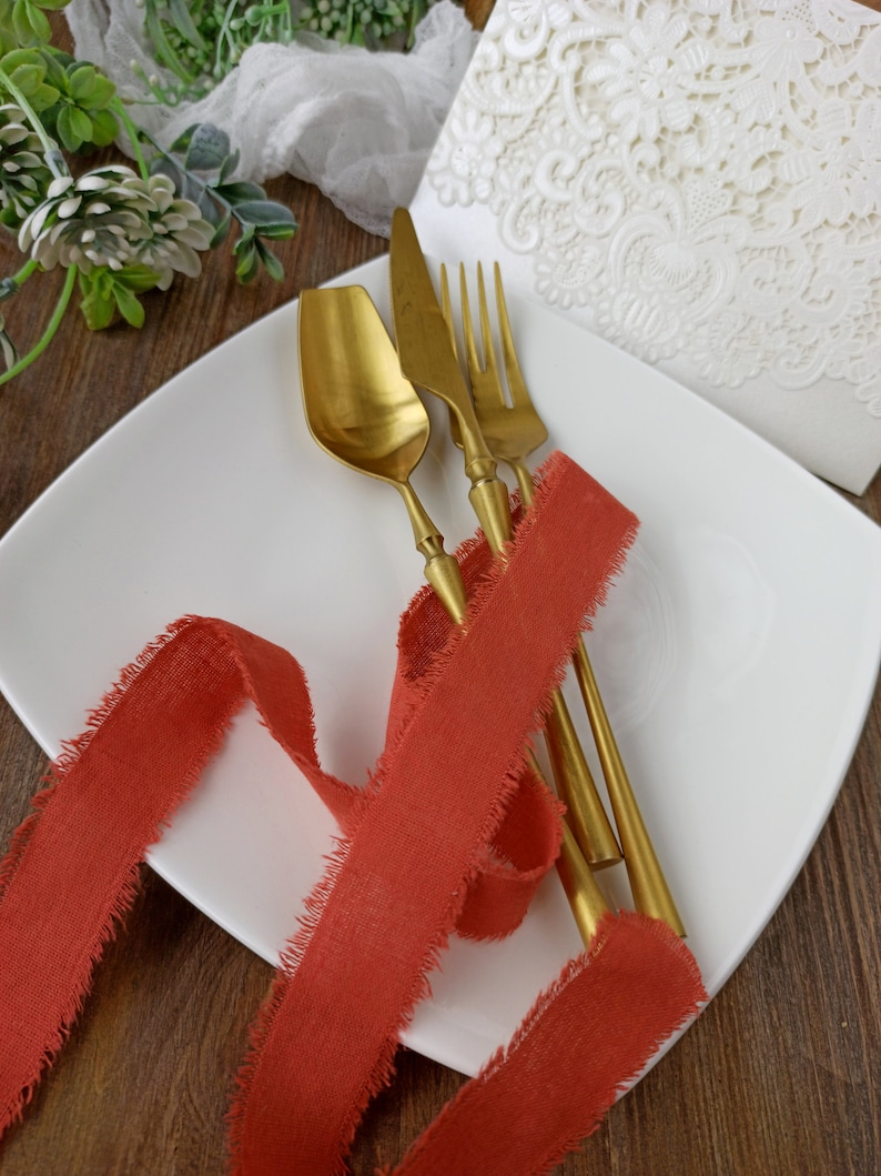 May include: A white plate with a red ribbon and gold cutlery. The ribbon has frayed edges and the cutlery is arranged on top of the ribbon.