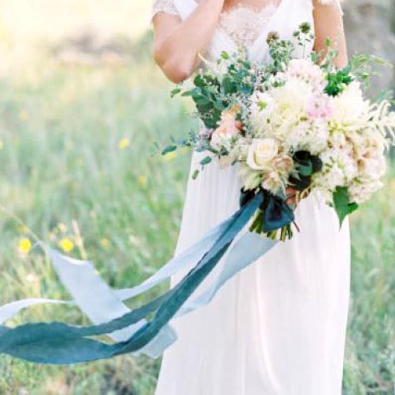 May include: A bride holds a bouquet of white and pink flowers with greenery and blue ribbon streamers.