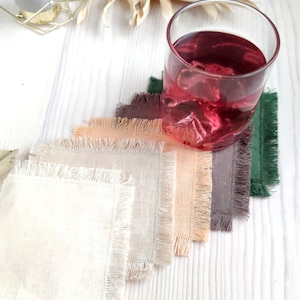 May include: Set of five square fabric coasters with frayed edges in various neutral colors, including beige, light brown, peach, and green. The coasters are arranged in a fan shape on a white wooden surface. A glass of pink liquid with ice cubes sits on top of the coasters.