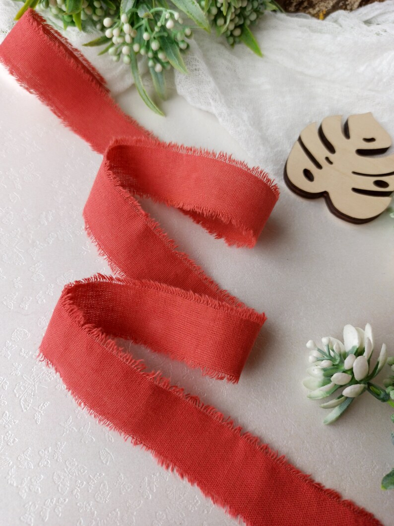 May include: A length of coral red frayed ribbon with a raw edge. The ribbon is folded and laid out on a white surface.