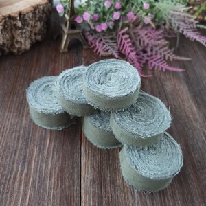 May include: A stack of six green fabric ribbon rolls, each with a frayed edge. The rolls are arranged in a pyramid shape on a wooden surface.