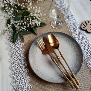 Peut inclure: Un gros plan sur une table rustique avec une nappe en dentelle blanche, un chemin de table en toile de jute et une assiette grise mouchet&eacute;e avec des couverts dor&eacute;s.