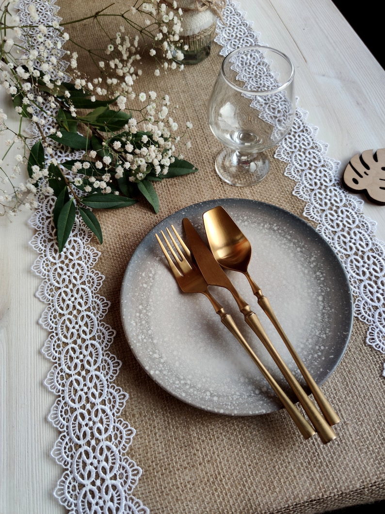 Peut inclure: Un gros plan sur une table rustique avec une nappe en dentelle blanche, un chemin de table en toile de jute et une assiette grise mouchet&eacute;e avec des couverts dor&eacute;s.