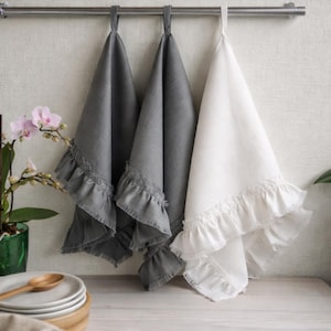 a set of three kitchen towels hanging on a rack, with two gray towels and one white towel. The towels have ruffled edges and appear to be made of a soft, textured fabric. In the background, there is a green plant and a stack of plates, suggesting a cozy and functional kitchen setting.