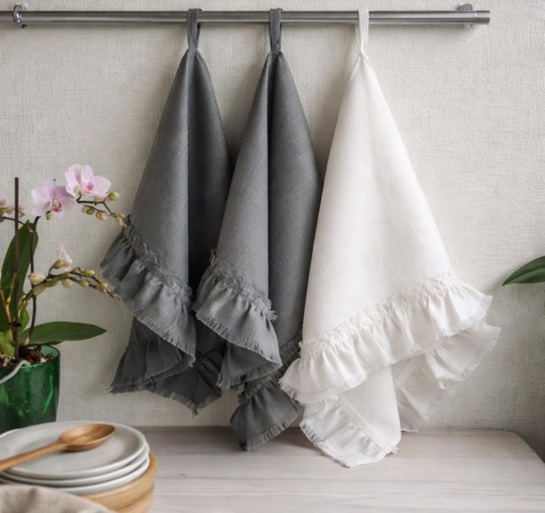 a set of three kitchen towels hanging on a rack, with two gray towels and one white towel. The towels have ruffled edges and appear to be made of a soft, textured fabric. In the background, there is a green plant and a stack of plates, suggesting a cozy and functional kitchen setting.