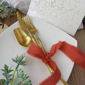 May include: A white plate with a gold fork, knife, and spoon tied together with a red ribbon. The plate is on a wooden surface with green plants in the background.