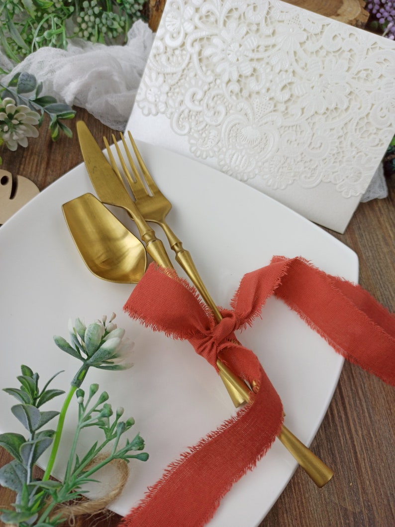 May include: A white plate with a gold fork, knife, and spoon tied together with a red ribbon. The plate is on a wooden surface with green plants in the background.