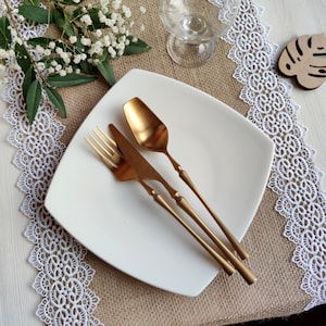 Peut inclure: Une assiette carr&eacute;e blanche avec une fourchette, un couteau et une cuill&egrave;re dor&eacute;s sur un chemin de table en toile de jute avec une bordure en dentelle blanche.