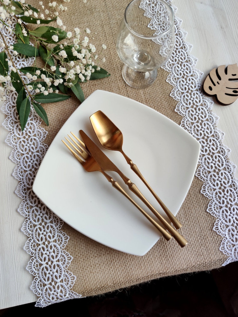 Peut inclure: Une assiette carr&eacute;e blanche avec une fourchette, un couteau et une cuill&egrave;re dor&eacute;s sur un chemin de table en toile de jute avec une bordure en dentelle blanche.