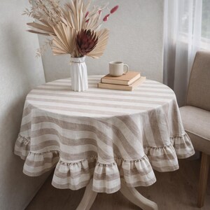 a cozy corner of a room with a round table covered in a striped tablecloth. On the table, there is a vase with dried flowers, a stack of books, and a cup of coffee.