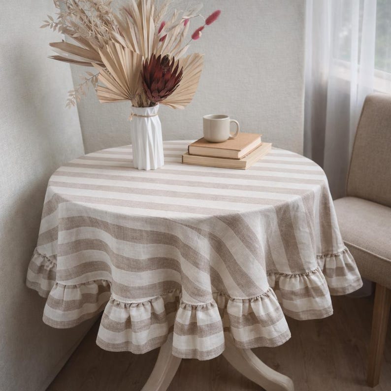 a cozy corner of a room with a round table covered in a striped tablecloth. On the table, there is a vase with dried flowers, a stack of books, and a cup of coffee.