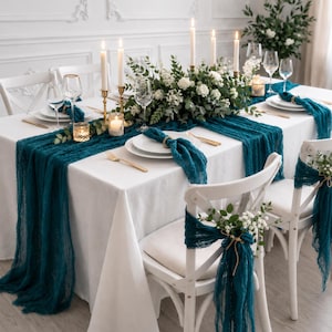May include: A beautifully set dining table with a white tablecloth, teal table runner, and matching chair sashes. The table is adorned with white plates, gold cutlery, candles, and a floral centerpiece, creating an elegant ambiance.