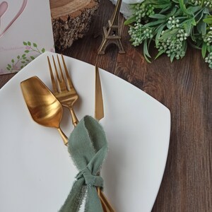 May include: A white square plate with a gold fork, knife, and spoon tied together with a sage green ribbon.