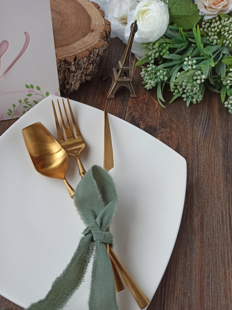 May include: A white square plate with a gold fork, knife, and spoon tied together with a sage green ribbon.