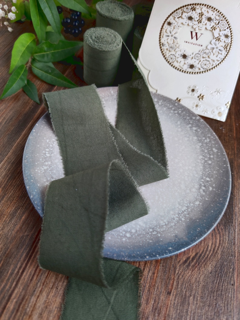 May include: A dark green fabric ribbon with frayed edges is displayed on a white speckled plate. The ribbon is long and wide, and it is folded in a way that shows off its texture. There are two rolls of the same fabric ribbon in the background.