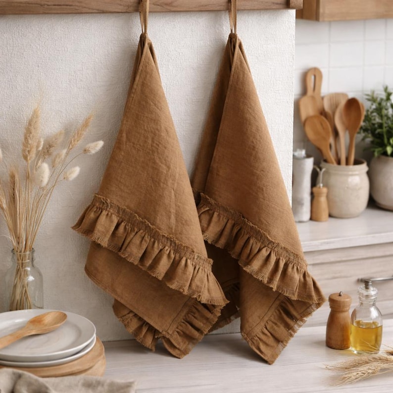 a kitchen counter with two brown ruffled towels hanging on a wooden rack. The towels have a rustic, natural appearance. On the counter, there are a few kitchen utensils, including a wooden spoon, a wooden pepper grinder, and a glass bottle containing a yellow liquid. The overall scene has a warm, cozy atmosphere.