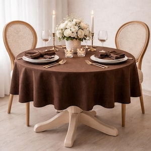 May include: A round table covered with a dark brown tablecloth, set for two with white plates, brown napkins, gold forks, and wine glasses. A white floral centerpiece in a textured vase sits between two tall candles and two small votive candles.