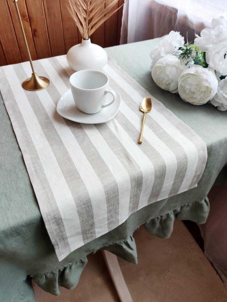 May include: A white linen table runner with brown stripes, placed on a green linen tablecloth with ruffles. A white cup and saucer, a gold spoon, and a vase with dried palm leaves are on the table runner.