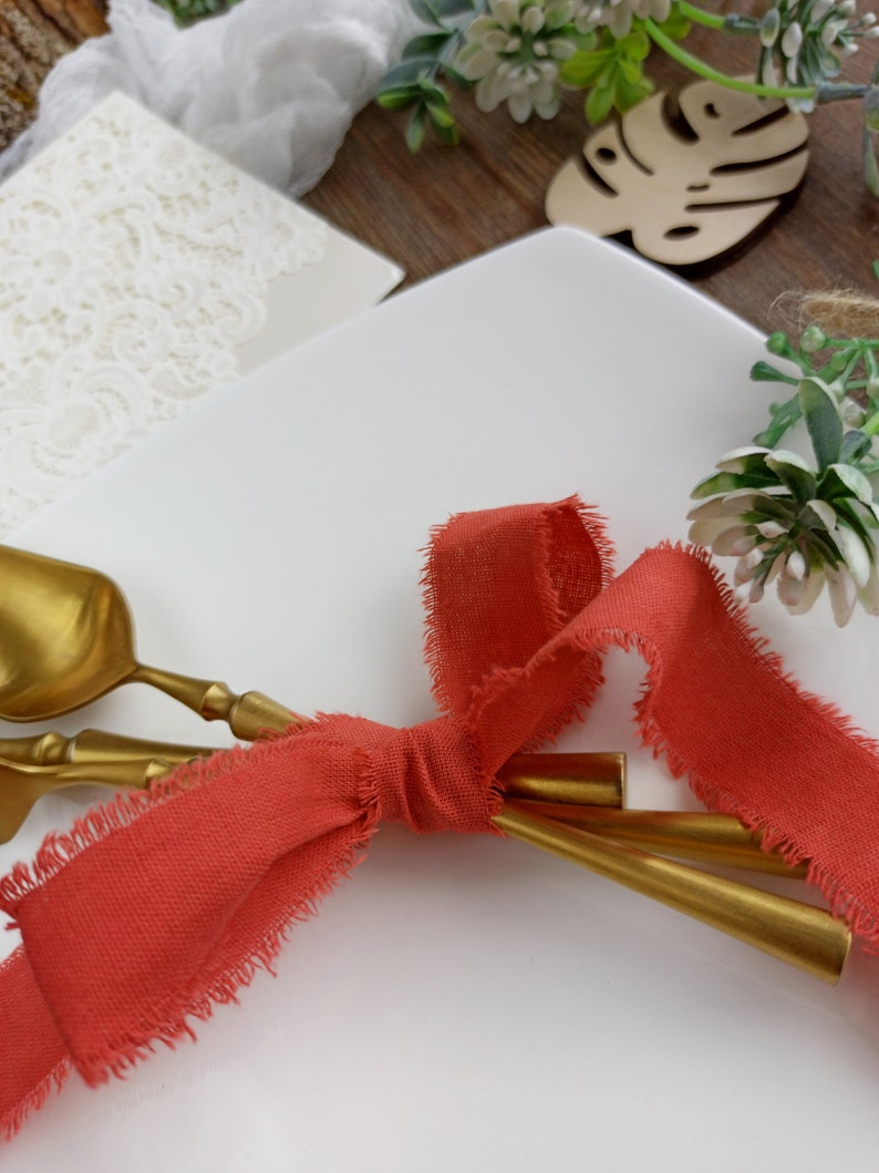 May include: Two gold utensils tied together with a coral red ribbon. The ribbon has frayed edges. The utensils are on a white plate.