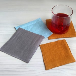 May include: A selection of square linen coasters in grey, light blue, and orange. A glass of red liquid rests on one of the orange coasters. The coasters are displayed on a white wooden surface.