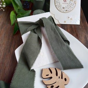 May include: A white invitation card with a gold floral design and the text "W Invitation". The card is tied with a dark green ribbon and placed on a white plate with a wooden leaf cutout.