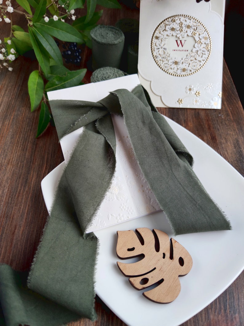 May include: A white invitation card with a gold floral design and the text "W Invitation". The card is tied with a dark green ribbon and placed on a white plate with a wooden leaf cutout.