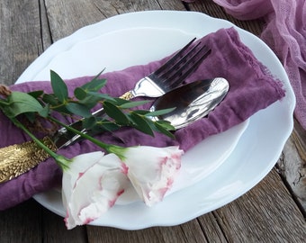 May include: A table setting with white plates, a purple linen napkin, and silver cutlery. The arrangement includes white flowers with pink edges and eucalyptus sprigs, set against a weathered wooden surface. The plates have a scalloped edge.