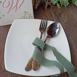 May include: A white square plate with a fork and spoon tied together with a sage green ribbon. The fork and spoon have gold-colored handles with a textured design.