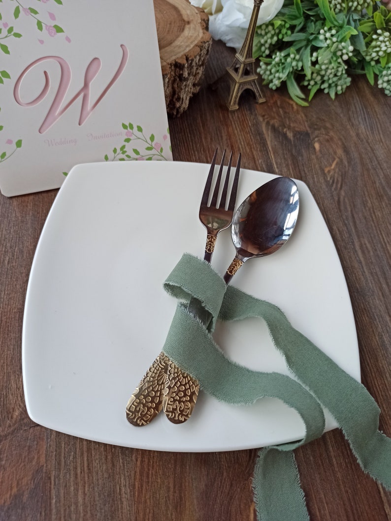 May include: A white square plate with a fork and spoon tied together with a sage green ribbon. The fork and spoon have gold-colored handles with a textured design.