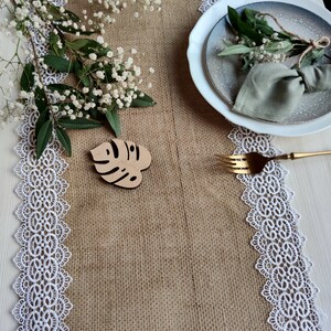 Peut inclure: Un chemin de table en toile de jute avec une bordure en dentelle blanche, un marque-place en bois en forme de feuille et une assiette blanche avec une serviette verte et de la verdure sur le dessus.