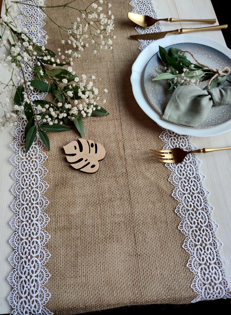 Peut inclure: Un chemin de table en toile de jute avec une bordure en dentelle blanche, un marque-place en bois en forme de feuille et une assiette blanche avec une serviette verte et de la verdure sur le dessus.