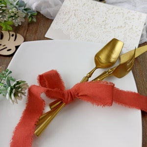 May include: Two gold-toned utensils tied together with a coral-colored ribbon on a white square plate. The ribbon is tied in a bow with frayed edges.