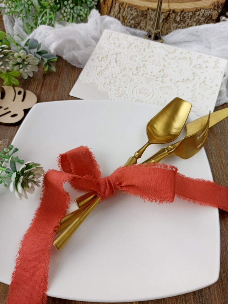 May include: Two gold-toned utensils tied together with a coral-colored ribbon on a white square plate. The ribbon is tied in a bow with frayed edges.