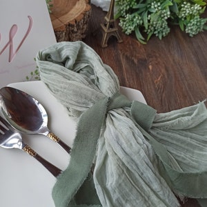 May include: A sage green fabric napkin with a frayed edge, tied with a sage green ribbon, and placed on a white plate with silver cutlery.