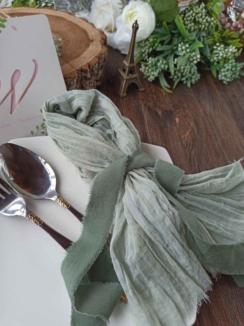 May include: A sage green fabric napkin with a frayed edge, tied with a sage green ribbon, and placed on a white plate with silver cutlery.