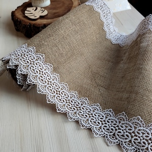 Peut inclure: Un chemin de table en toile de jute brune avec une bordure en dentelle blanche. Le chemin de table est pli&eacute; en deux et la bordure en dentelle est visible des deux c&ocirc;t&eacute;s.