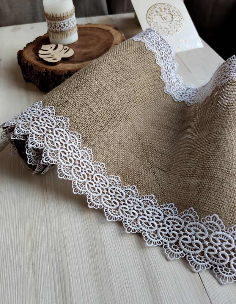 Peut inclure: Un chemin de table en toile de jute brune avec une bordure en dentelle blanche. Le chemin de table est pli&eacute; en deux et la bordure en dentelle est visible des deux c&ocirc;t&eacute;s.