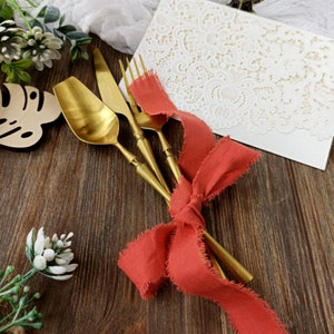 May include: Gold cutlery set with a red ribbon tied around the handles. The cutlery is on a wooden surface with a white lace overlay and a small potted plant.