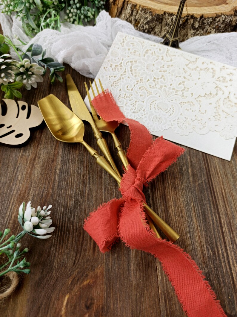 May include: Gold cutlery set with a red ribbon tied around the handles. The cutlery is on a wooden surface with a white lace overlay and a small potted plant.