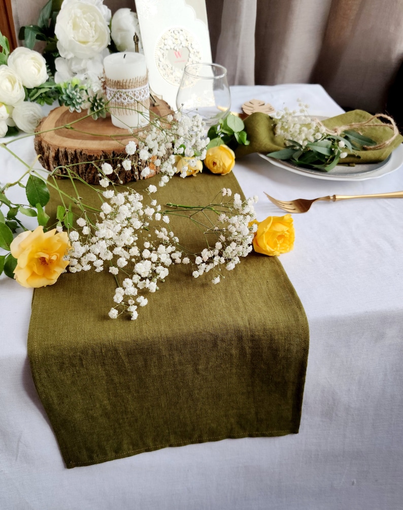 May include: A green linen table runner with white baby's breath and yellow roses. The runner is on a white tablecloth and there is a place setting with a white plate, a gold fork, and a green linen napkin.