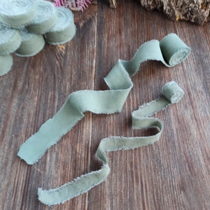 May include: Two rolls of sage green fabric ribbon with frayed edges. The ribbon is laid out on a wooden surface.