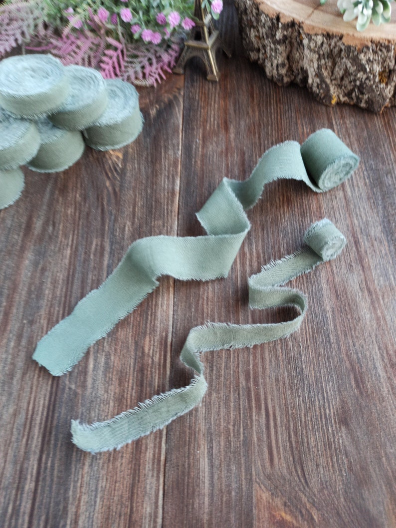May include: Two rolls of sage green fabric ribbon with frayed edges. The ribbon is laid out on a wooden surface.