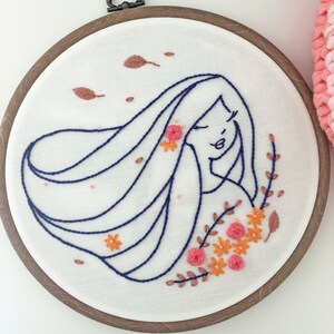 Könnte beinhalten: Stickbild mit Stickerei, das eine Frau mit langen, fließenden Haaren und floralen Details in Rosa-, Orange- und Brauntönen zeigt.
