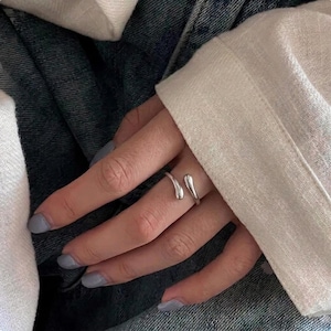 May include: A silver-coloured open ring featuring two smooth, teardrop-shaped ends that face each other. The ring has a polished, reflective metallic finish and a simple, minimalist band design that wraps partially around the finger.