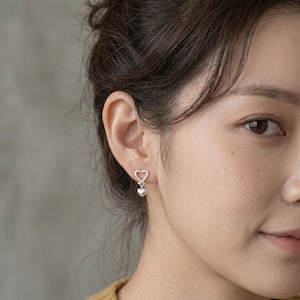 May include: A silver stud earring featuring an open heart-shaped frame with a small, solid heart charm dangling from the bottom. The earring is crafted from polished silver metal and is worn in the earlobe, showcasing a delicate and refined design.