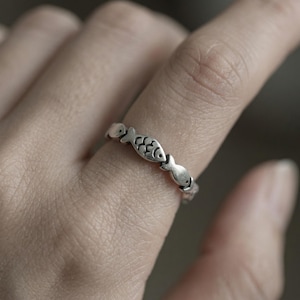 May include: A silver ring featuring a repeating fish design. The ring is worn on a finger, showcasing the detailed fish silhouettes. The ring's design is simple yet elegant, with each fish carefully crafted.