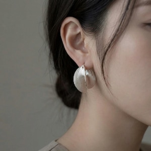 May include: Silver hoop earrings with a unique, layered design. Each earring features a half-circle shape with a textured, ridged surface, creating a fan-like appearance. The earrings are shown on a person's ear, highlighting their elegant and modern style.