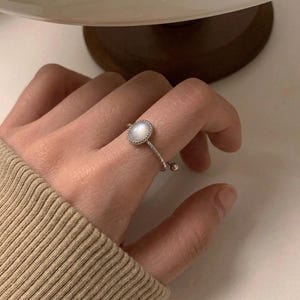May include: A silver-toned ring featuring a small, round, iridescent white stone set in a beaded bezel. The band has a textured, twisted appearance and includes a small, dangling silver-toned bead charm attached to the side of the ring setting.
